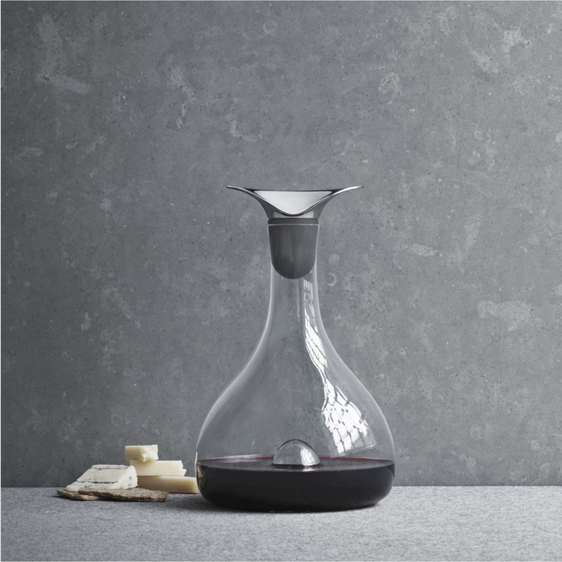 The Georg Jensen Wine & Bar Carafe, filled with red wine, sits beside cheese and crackers on a grey surface. A textured grey wall in the background creates an elegant setting for enjoying perfectly aerated wine.