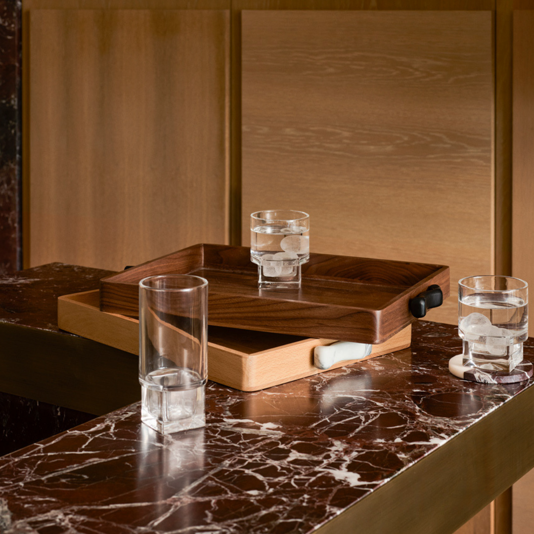 The Greg Natale Terreno Rectangle Marble Handled Tray with a black handle sits on a brown marble countertop, holding a glass of iced water. Two more glasses with ice are beside the tray, set against light wood paneling in the background.