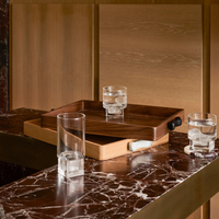 The Greg Natale Terreno Rectangle Marble Handled Tray with a black handle sits on a brown marble countertop, holding a glass of iced water. Two more glasses with ice are beside the tray, set against light wood paneling in the background.