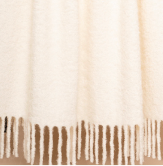 A close-up of the Brushed Boucle Throw by Thread & Butter, featuring a cream-colored, soft, textured finish with a natural sheen and long, twisted fringe along the bottom edge.