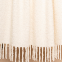 A close-up of the Brushed Boucle Throw by Thread & Butter, featuring a cream-colored, soft, textured finish with a natural sheen and long, twisted fringe along the bottom edge.