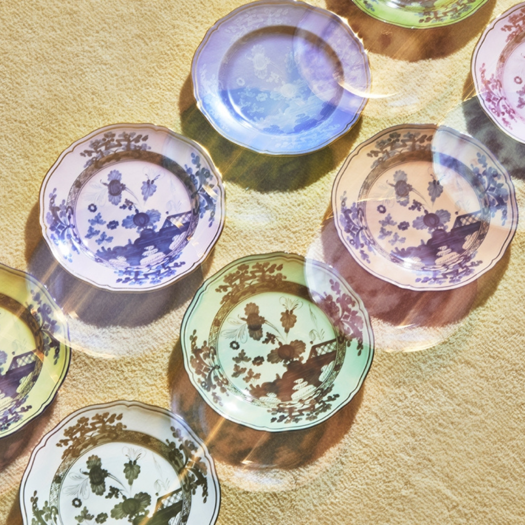 Several Ginori 1735 Oriente Italiano Dinnerware Bario plates with chinoiserie floral designs are arranged on a textured beige surface, while colorful light reflections cast a rainbow effect across the scene.