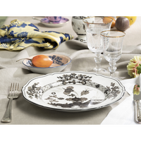 A table set with a Ginori 1735 Oriente Italiano Dinnerware Albus plate, two ornate glasses, cutlery, napkin, and a small dish of yellow fruit; a flower, patterned cloth, and extra glassware can be seen in the background.