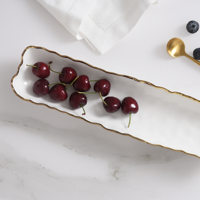 The Portofino Small Tray by PAMPA BAY, a rectangular porcelain serving dish with gold trim, holds nine cherries. Nearby on the white marble surface are a gold spoon, blueberries, and a folded white napkin.