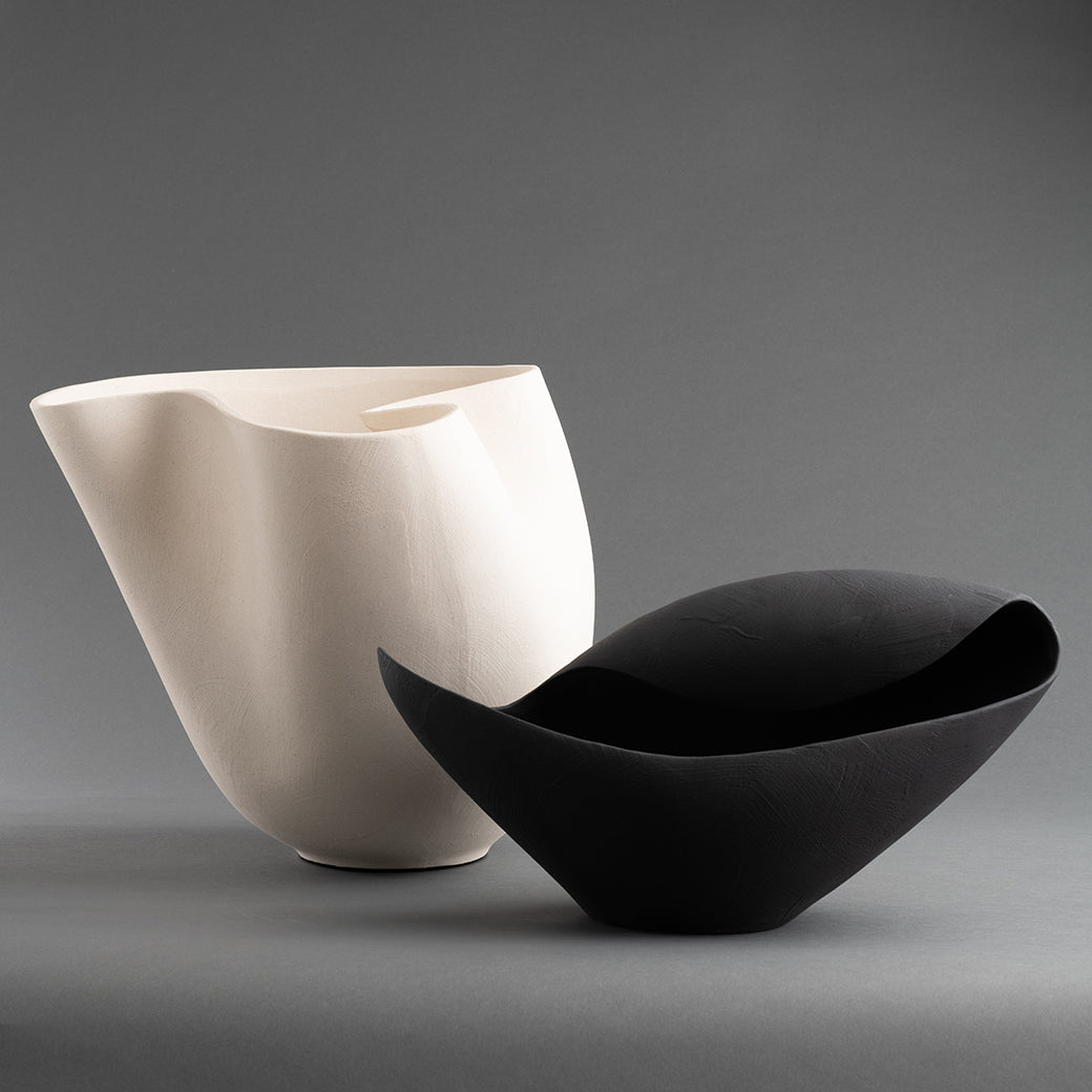 A white Carmen Vase by FlowDecor Inc. with an abstract silhouette stands next to a black sculptural bowl against a grey background.