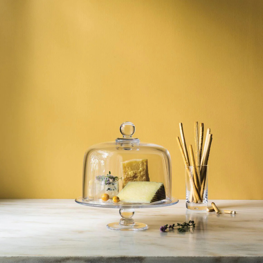 A Simon Pearce Hartland Cakeplate with a glass cloche displays cheese wedges, herbs, and golden berries on marble, while a nearby glass holds crisp breadsticks—all set before a warm yellow wall.