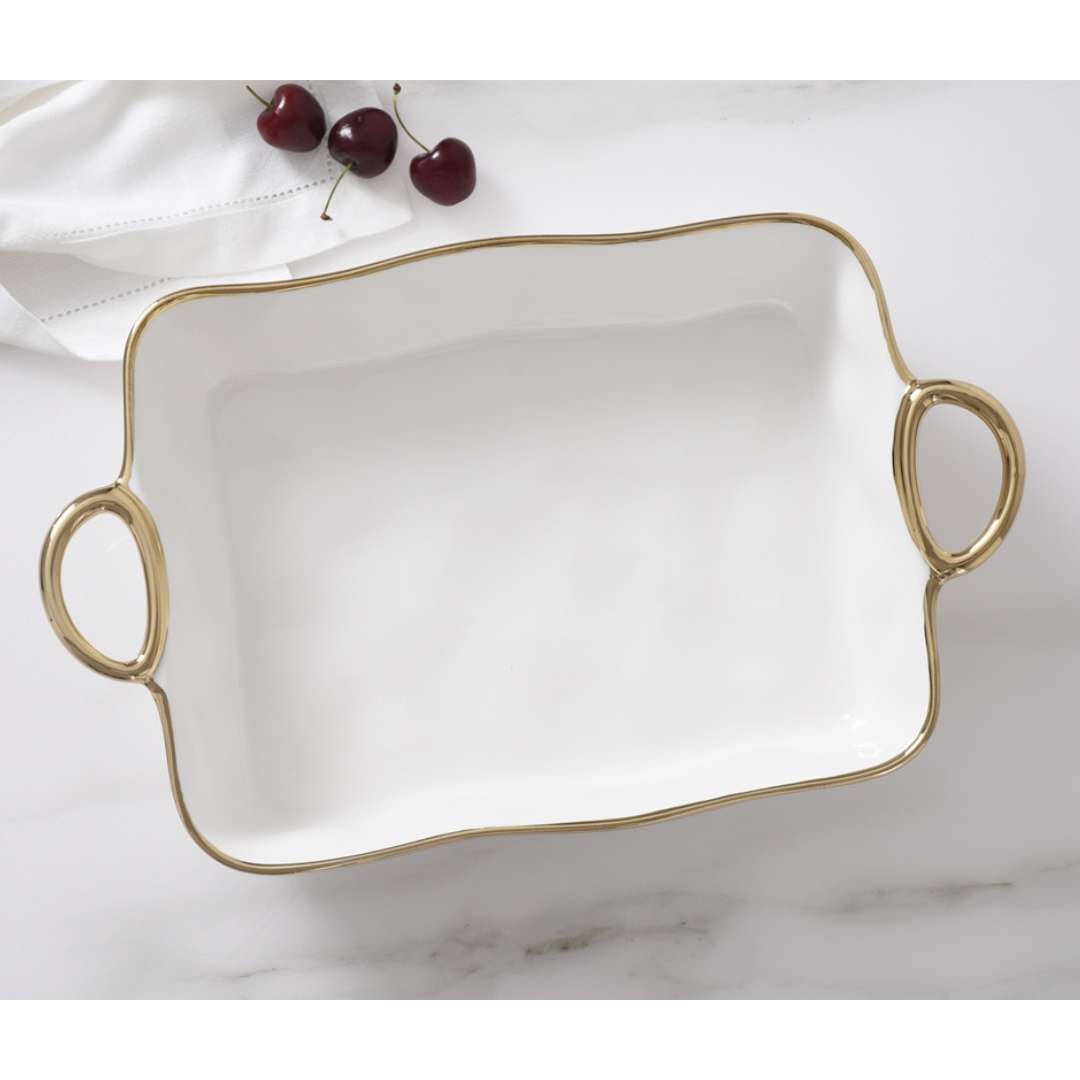 The PAMPA BAY Golden Handle Rectangle Baking Dish, a white porcelain piece with gold handles and trim, sits on a marble surface. Three cherries and a folded white cloth are in the background.