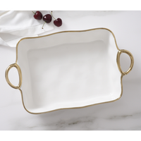 The PAMPA BAY Golden Handle Rectangle Baking Dish, a white porcelain piece with gold handles and trim, sits on a marble surface. Three cherries and a folded white cloth are in the background.