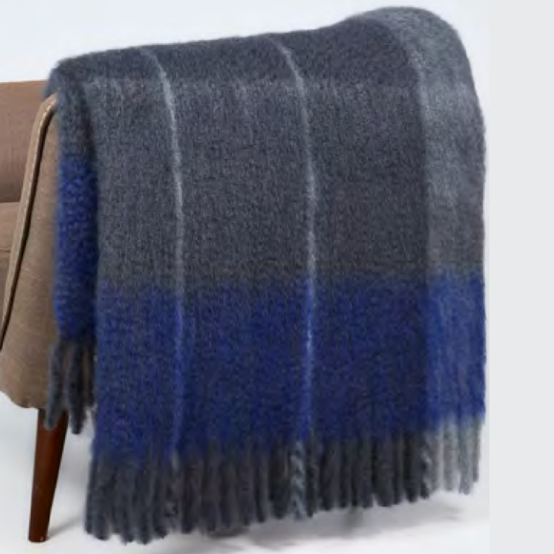 The Mantas Ezcaray Twist Frame Throw, a fuzzy mohair blanket handcrafted in Spain with blue and gray plaid and fringed edges, is draped over a brown chair.