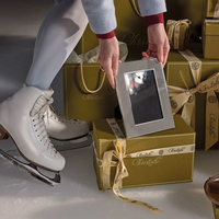 A person in white ice skates holds the America Stainless Steel Frame by Christofle above a stack of gold gift boxes tied with cream-colored Christofle ribbons.