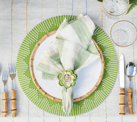 A table is set with a Kim Seybert Dream Weaver Placemat in green & white, a white plate with bamboo rim, plaid napkin in crocheted flower ring, bamboo-handled cutlery, and glasses on a striped tablecloth.