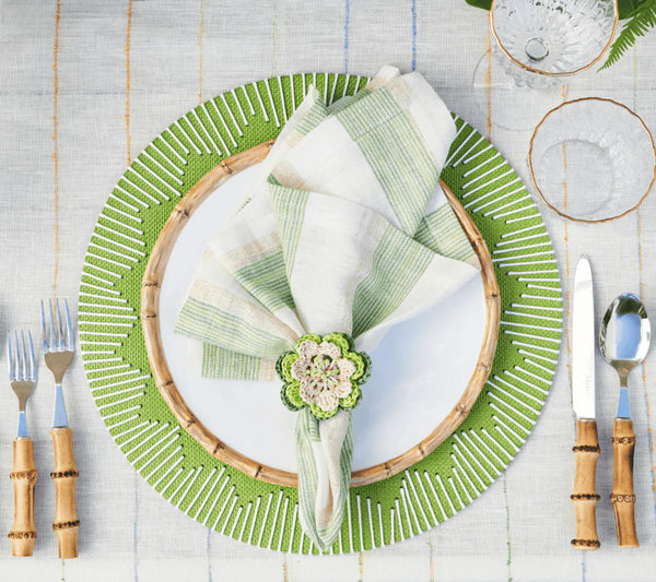 A table is set with a Kim Seybert Dream Weaver Placemat in green & white, a white plate with bamboo rim, plaid napkin in crocheted flower ring, bamboo-handled cutlery, and glasses on a striped tablecloth.