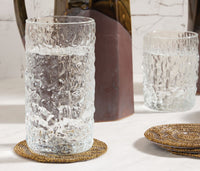 A glass from the Blue Pheasant Frederick Glassware Set of 6 - Clear, filled with water, rests on a beaded coaster on marble, with another matching glass and a brown ceramic pitcher in the background.