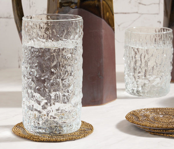 A glass from the Blue Pheasant Frederick Glassware Set of 6 - Clear, filled with water, rests on a beaded coaster on marble, with another matching glass and a brown ceramic pitcher in the background.