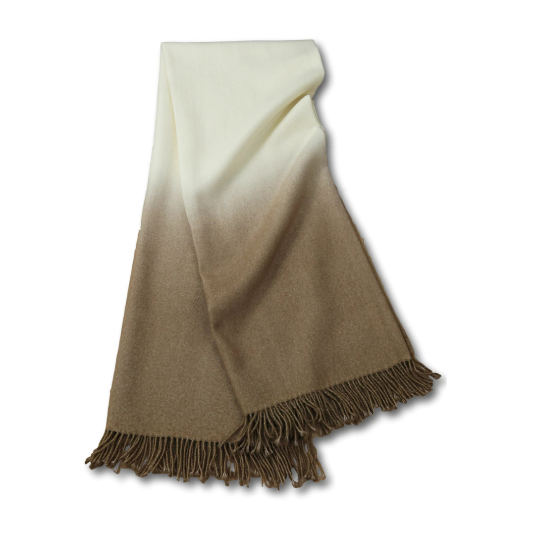 The Johanna Howard Dip Dye Throw is a soft baby alpaca blanket with white fading into light and dark brown shades, featuring fringed edges. Shown neatly folded on a white background.