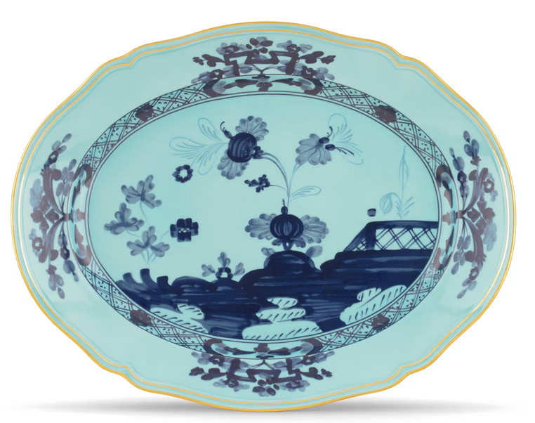 The Oriente Italiano Dinnerware Iris by Ginori 1735 is an oval Italian porcelain plate with a scalloped edge, featuring chinoiserie floral and landscape patterns in blue with gold accents on a light blue background.