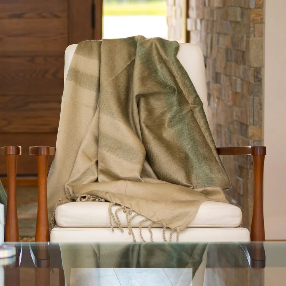 A Purple Nest Kallpa Alpaca Throw - Tobacco Olive & Khaki with green and tan stripes and fringe draped over a white armchair.