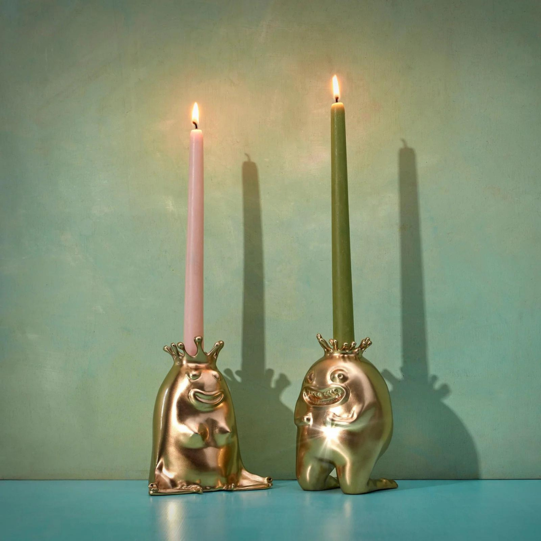 The L’Objet Haas King & Queen Candlesticks hold pink and green taper candles, both lit, against a plain teal background—luxury collectibles that cast crisp shadows on the wall.