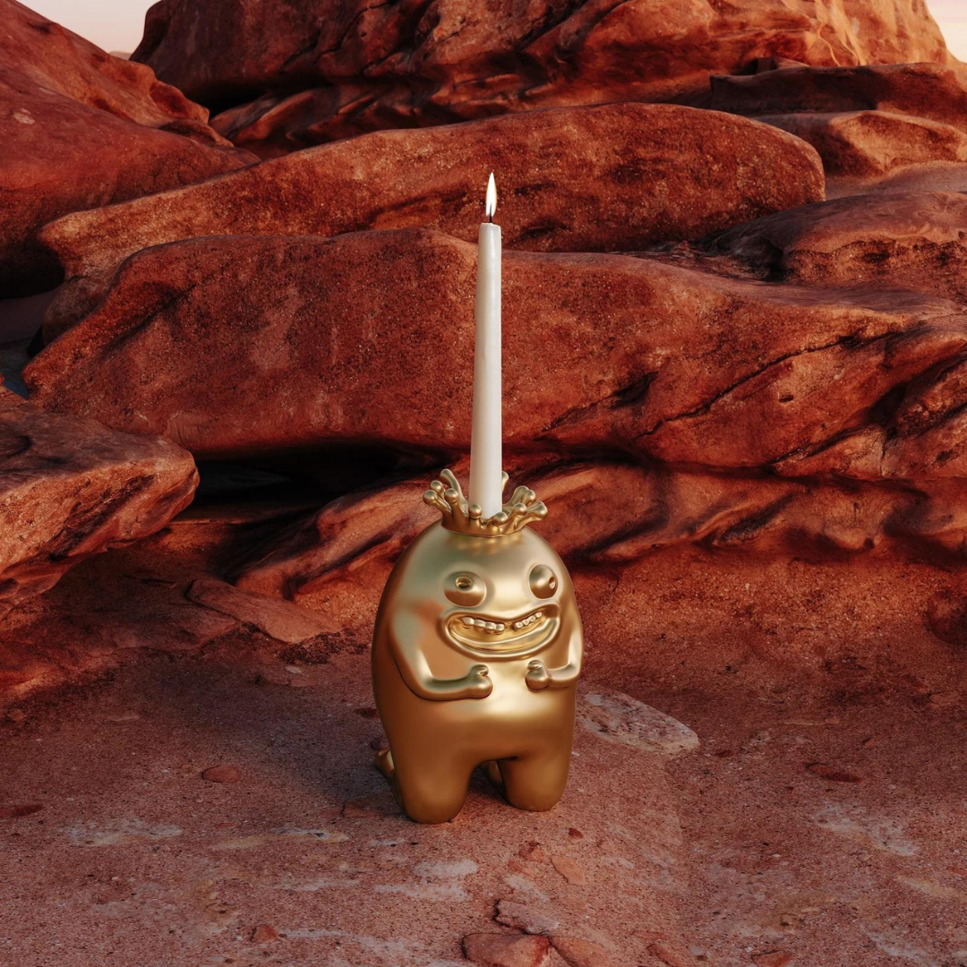 The Haas King & Queen Candlesticks by L’Objet feature whimsical, gold-toned designs and are shown with a white candle atop reddish desert rocks—an ideal luxury collectible for those who love unique decor.
