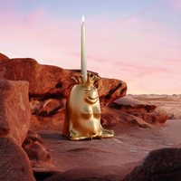 A gold Haas King & Queen Candlestick by L’Objet, shaped like a crowned frog, holds a white candle amid reddish-brown rocks under a pink and blue desert sky—an exquisite luxury collectible.
