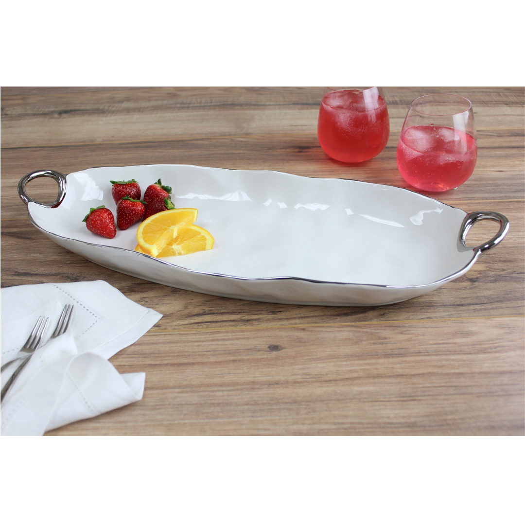The PAMPA BAY Handle Deep Serving Platter Silver, a white porcelain tray with silver titanium trim and handles, holds strawberries and orange slices on a wooden table beside pink drinks, napkins, and forks.