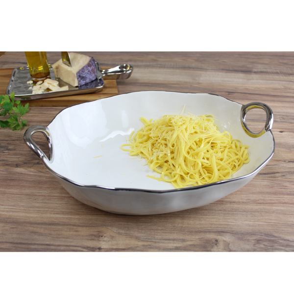 The PAMPA BAY Handle Deep Oval Serving Bowl Silver, made of white porcelain with a silver trim, holds uncooked pasta. In the background, a cutting board features cheese, oil, and chopped garlic on a wooden surface.