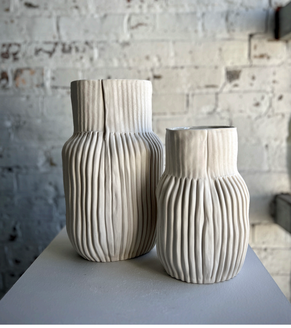 Two cream Series 02 Bottle Vases by CYM Warkov Ceramics, featuring vertical ridges and a modern handcrafted look, sit on a white surface before a textured white brick wall. The vases differ in height for added visual interest.