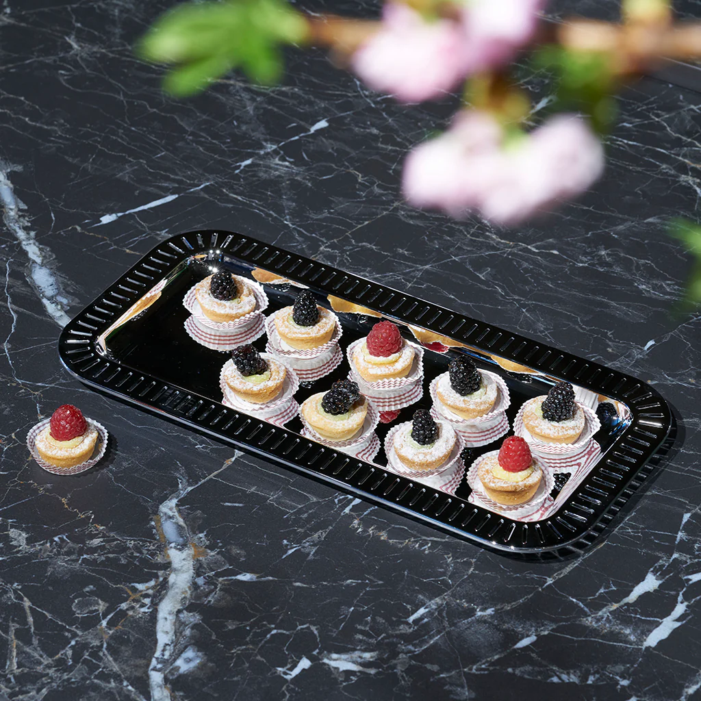 The Anna New York Striations Silver Small Rectangle Serving Tray holds fruit-topped pastries in paper cups on a dark marble surface, with one pastry beside the tray and blurred pink flowers and green leaves in the foreground.