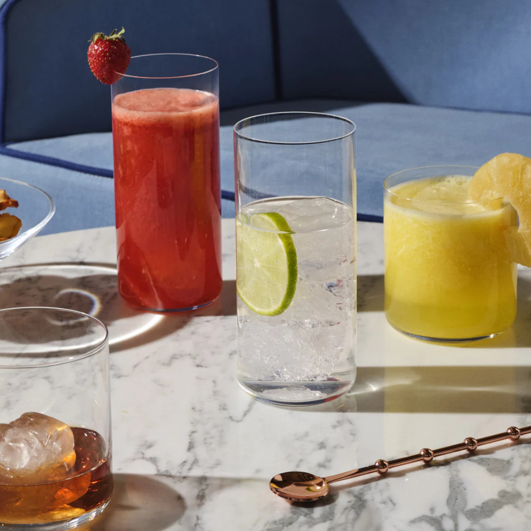 Four drinks on a marble table in Nude Glassware's Finesse Barware Set of 4 lead-free crystal whisky glasses: red with strawberry, clear with lime, yellow with lemon, and amber with ice. A gold spoon sits in front; a blue sofa completes the scene.