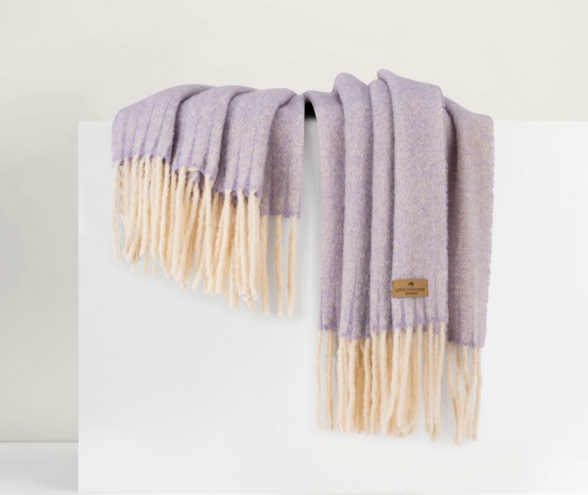 The Andes Throw by Lands Downunder, made of soft lavender baby alpaca with long cream fringes and a small brown label, is draped over a white surface against a neutral, minimalistic background.