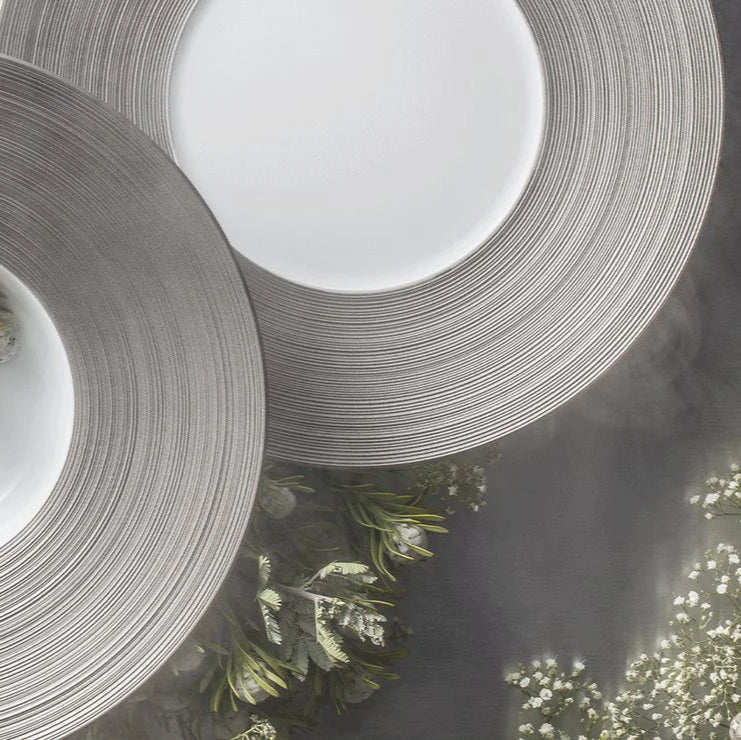 Two modern JL Coquet Hemisphere Dinnerware Platinum plates featuring wide, textured platinum rims overlap on a gray tablecloth next to greenery and white flowers, creating a sophisticated table setting.