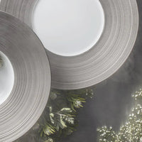 Two modern JL Coquet Hemisphere Dinnerware Platinum plates featuring wide, textured platinum rims overlap on a gray tablecloth next to greenery and white flowers, creating a sophisticated table setting.
