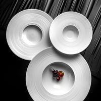 Three round, textured JL Coquet Hemisphere Dinnerware Satin White plates overlap on a black surface; berries and a small apple sit on the engraved bottom plate, while the other two remain empty.