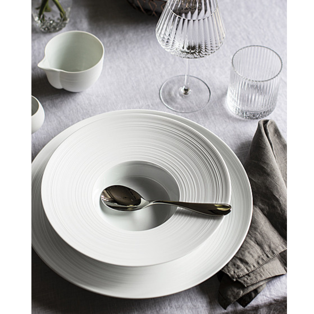 A table setting features a JL Coquet Hemisphere Dinnerware Satin White plate, a shiny spoon, gray napkin, clear wine and water glasses, and a small white creamer on a light gray tablecloth.