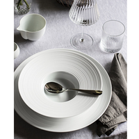 A table setting features a JL Coquet Hemisphere Dinnerware Satin White plate, a shiny spoon, gray napkin, clear wine and water glasses, and a small white creamer on a light gray tablecloth.