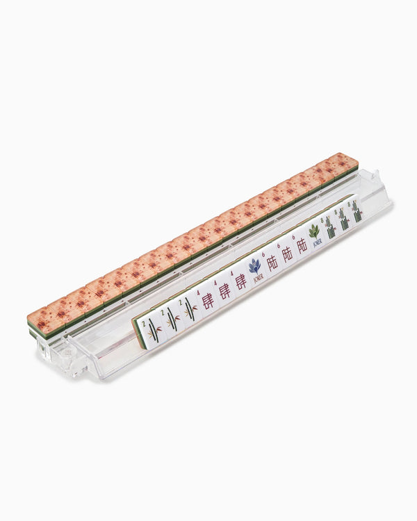 A clear plastic rack holds a row of Oh My Mahjong’s Mahjong Tiles Set - Heritage, showcasing bamboo and character designs with an heirloom-quality finish, accented by a pink floral-patterned strip along the top edge—a striking detail in this modern set.