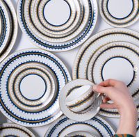 A hand holds a white Marie Daâge Rafia Stripes teacup and saucer with blue and gold accents, hand painted Limoges porcelain. The luxury tableware, by Marie Daâge, rests on a stack of matching plates.