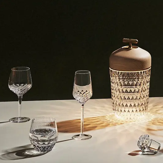 Three mouthblown Folia Stemware crystal glasses by Saint-Louis/Hermès and a hand-cut crystal ice bucket with a wooden lid from the Folia collection are arranged on a table. The ice bucket casts a decorative light pattern on the surface, while the dark background highlights the exquisite glassware.