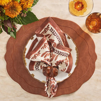 A fall table setting features the Kim Seybert Tailored Placemat Brown Set of 4, a gold-trimmed white plate, a patterned napkin in a ring, orange glassware, and a bouquet of orange flowers.