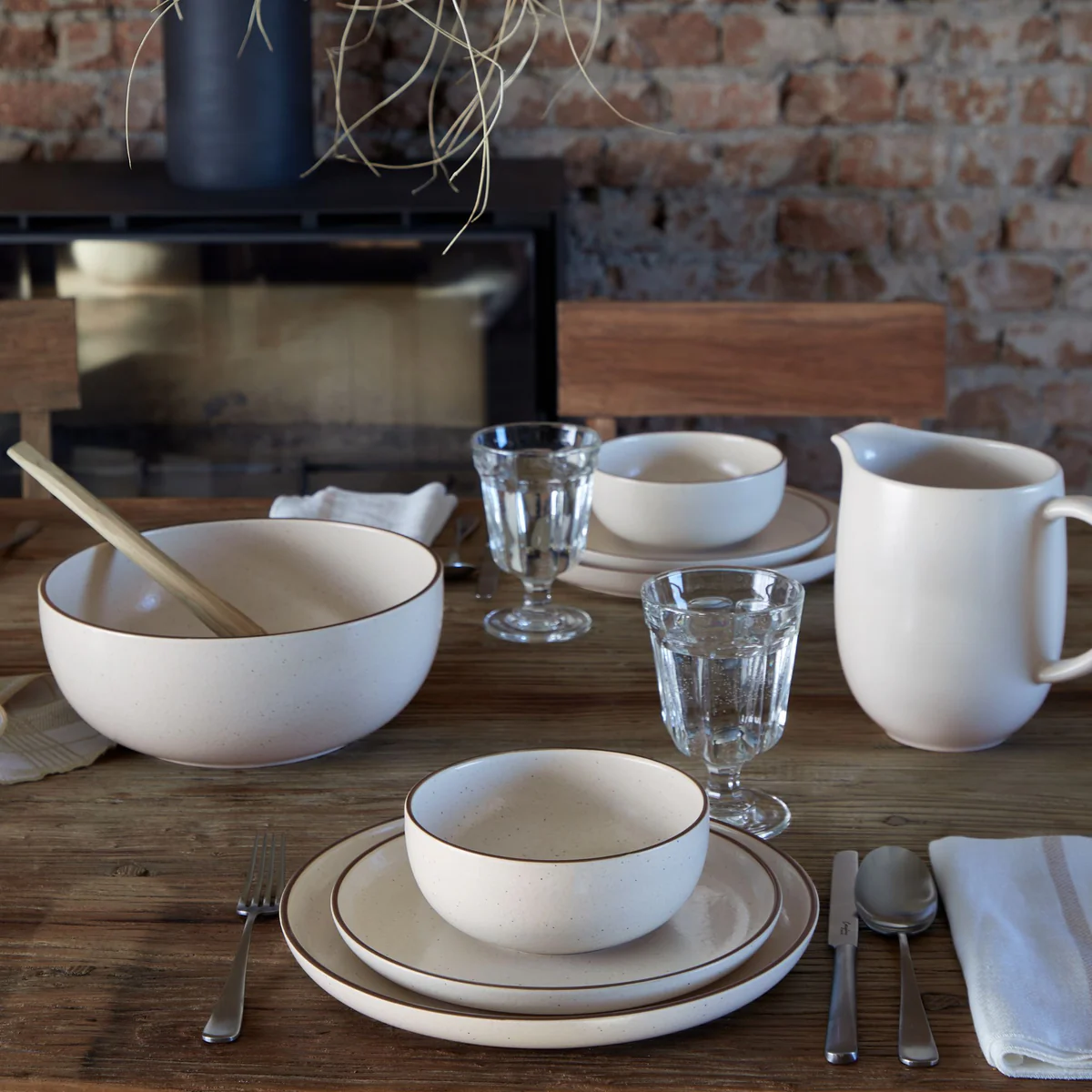 A rustic wooden table is set with Casafina/Costa Nova Pacifica Dinnerware Monterosa Beige, a pitcher, glasses, cutlery, and a large bowl with wooden spoon and napkin. Exposed brick wall and fireplace are in the background.