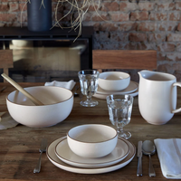 A rustic wooden table is set with Casafina/Costa Nova Pacifica Dinnerware Monterosa Beige, a pitcher, glasses, cutlery, and a large bowl with wooden spoon and napkin. Exposed brick wall and fireplace are in the background.