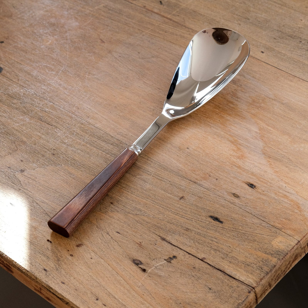 The Sabre Nature Rice Spoon, featuring a shiny metal finish and a colored wood handle, rests on a rustic wooden table as sunlight casts soft shadows across the surface.