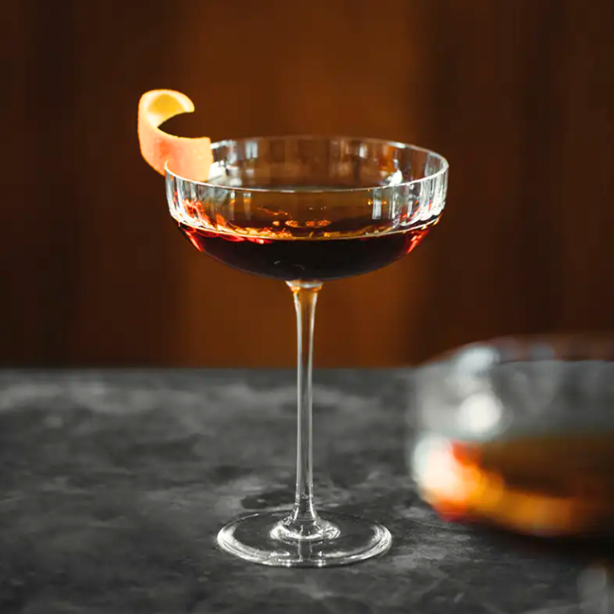 A Richard Brendon Fluted Coupe filled with a dark cocktail and topped with a curled orange peel garnish sits on a dark marble surface against a blurred brown background.