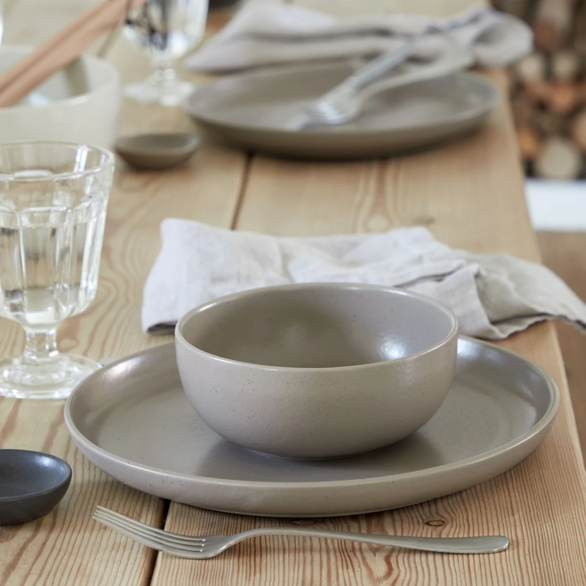 A calm, minimalist table features Casafina/Costa Nova’s Pacifica Chestnut set: a gray ceramic bowl on a matching plate, a glass of water, fork, cloth napkin, and an additional place setting in the background.