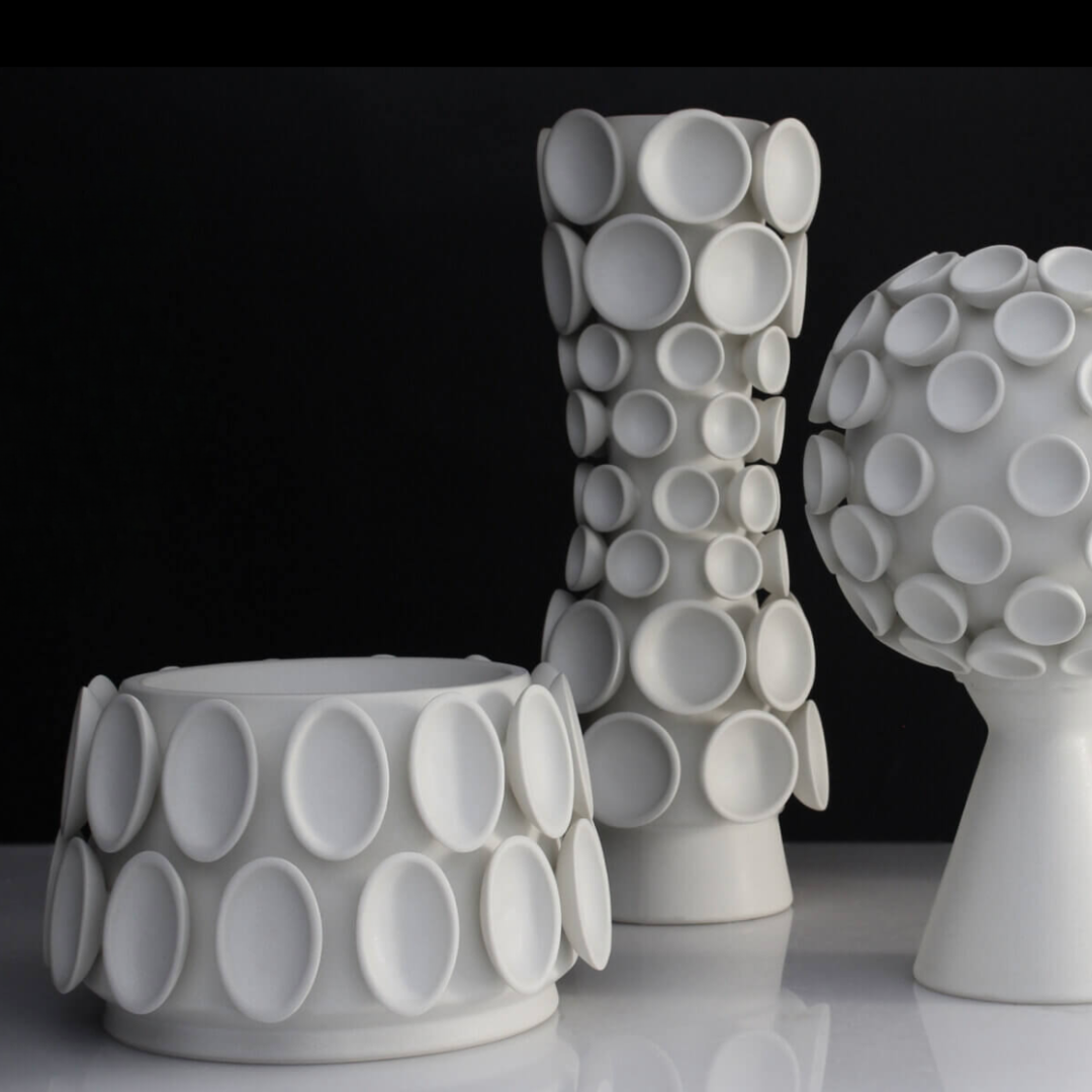 The Cumulus Collection Alabaster by KleinReid features three white ceramic pieces—a minimalist vase and a round bowl—each decorated with raised circular discs and modern textured designs, showcasing the refined elegance of glazed porcelain.