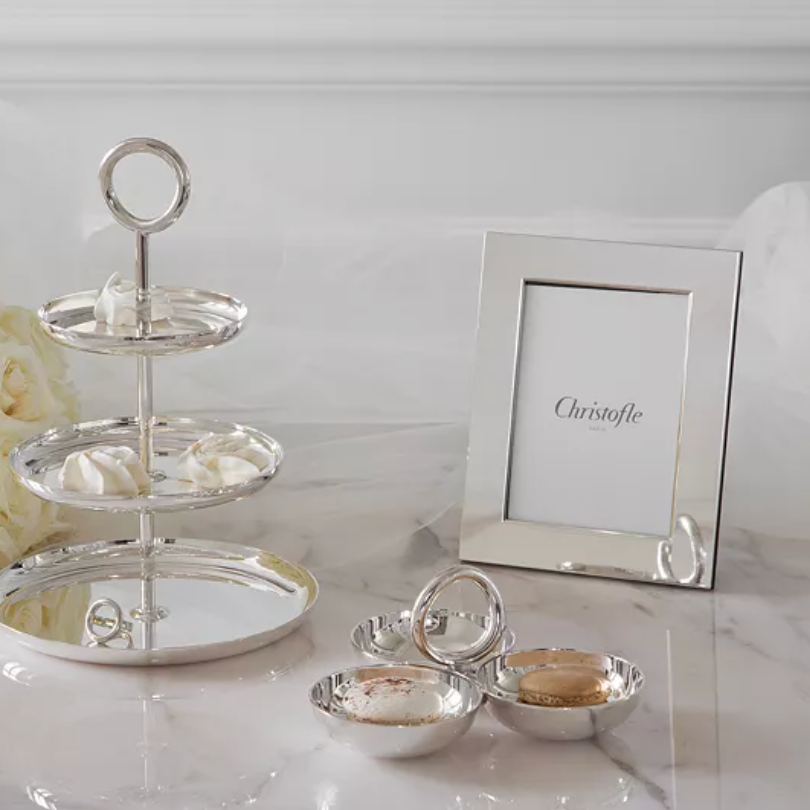 A three-tiered silver serving tray with white meringues, a Christofle Fidelio Silver Plated Frame 7x9.5, and a silver dish of macarons rest on marble, with white roses in the background.