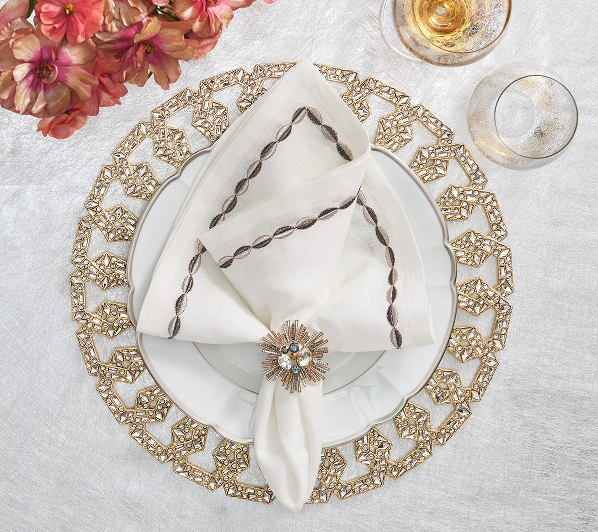 Create an elegant table setting with Kim Seybert's Swag Napkin Gunmetal & Silver Set of 4—white plate, gold placemat, jeweled napkin ring, glassware, and pink flowers on a white cloth—perfect for stylish holiday dining.
