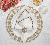 Create an elegant table setting with Kim Seybert's Swag Napkin Gunmetal & Silver Set of 4—white plate, gold placemat, jeweled napkin ring, glassware, and pink flowers on a white cloth—perfect for stylish holiday dining.
