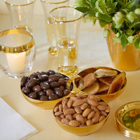 The Aerin Sintra 3-Bowl Server holds chocolate-covered nuts, plain almonds, and small round crackers. Nearby are gold-rimmed glasses, a gold-rimmed candle, a white napkin, and a green floral arrangement in a gold vase.