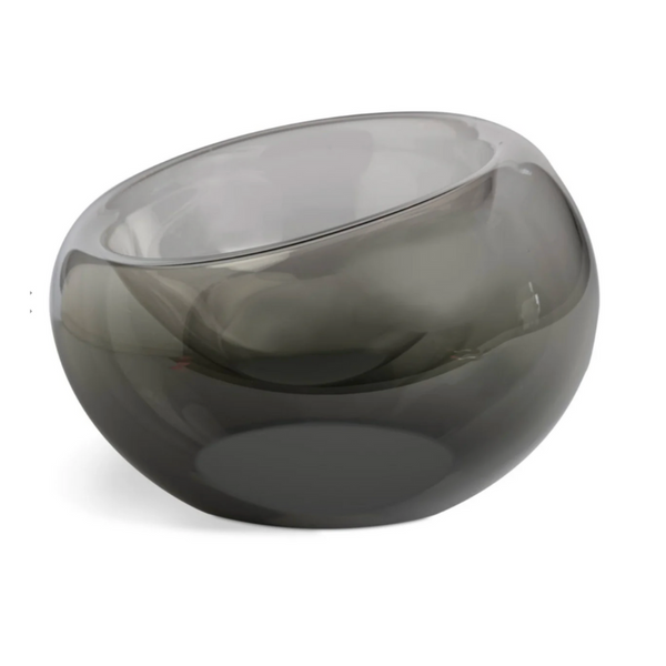 The Axiom Glass Caldera Vessel Medium 9" is a hand-blown, smoky gray glass centerpiece with a round shape and asymmetrical slanted opening, elegantly displayed on a white background.